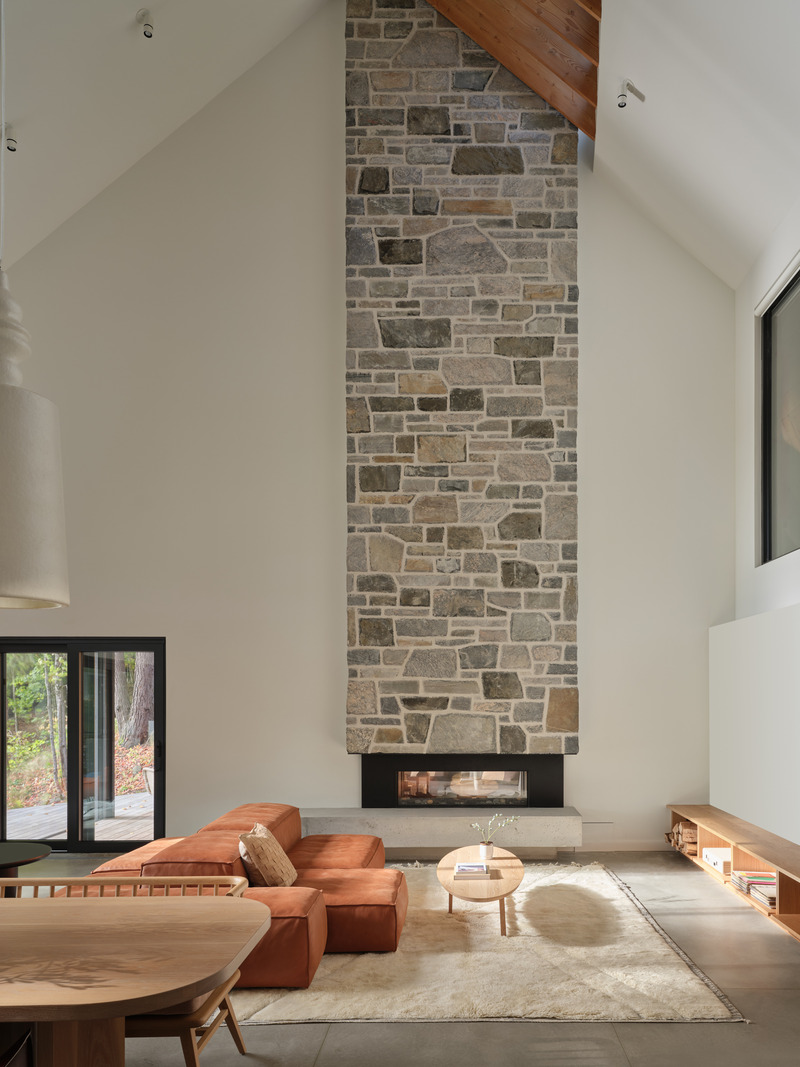 Press kit | 7600-02 - Press release | Shield and Shelter: An Embracing Lakeside Family Retreat that Opens Intimately and Sensitively to its Untamed Surroundings in the Heart of the Muskoka Region - Barbora Vokac Taylor Architect Inc. (BVT A) - Residential Architecture -  The living room features a double-sided fireplace, with a grand stone chimney adding aesthetic beauty as it rises through the levels of the structure to the exterior. From the exterior: the chimney marks the division between the house proper and the outdoor Muskoka Room.  - Photo credit: Doublespace
