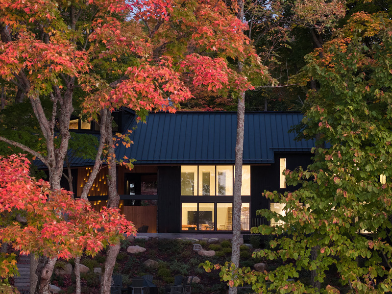 Press kit | 7600-02 - Press release | Shield and Shelter: An Embracing Lakeside Family Retreat that Opens Intimately and Sensitively to its Untamed Surroundings in the Heart of the Muskoka Region - Barbora Vokac Taylor Architect Inc. (BVT A) - Residential Architecture -   View from the lake: By day, the angular structure provides deliberate contrast to its organic setting, allowing for visitors to read that landscape against the neutral silhouette while carefully positioned "viewfinders" frame specific natural moments: Some bathe the interior in sunlight, other frame specific trees, and some overlook sunrises, grounding the home in its rugged setting.   - Photo credit: Doublespace