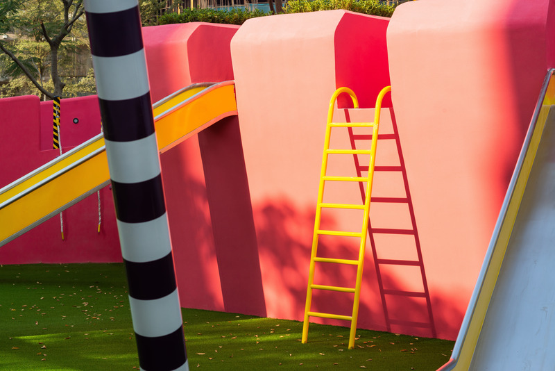 Dossier de presse | 5086-04 - Communiqué de presse | Les Résidences Buji par BAM : couleurs vives - Ballistic Architecture Machine - Architecture de paysage - Détail de la zone de toboggans aux Résidences Buji, où une échelle et un toboggan jaunes contrastent avec la paroi courbe rose pâle et la colonne rayée noir et blanc, sur une pelouse verte. - Crédit photo : Amey Kandalgaonkar