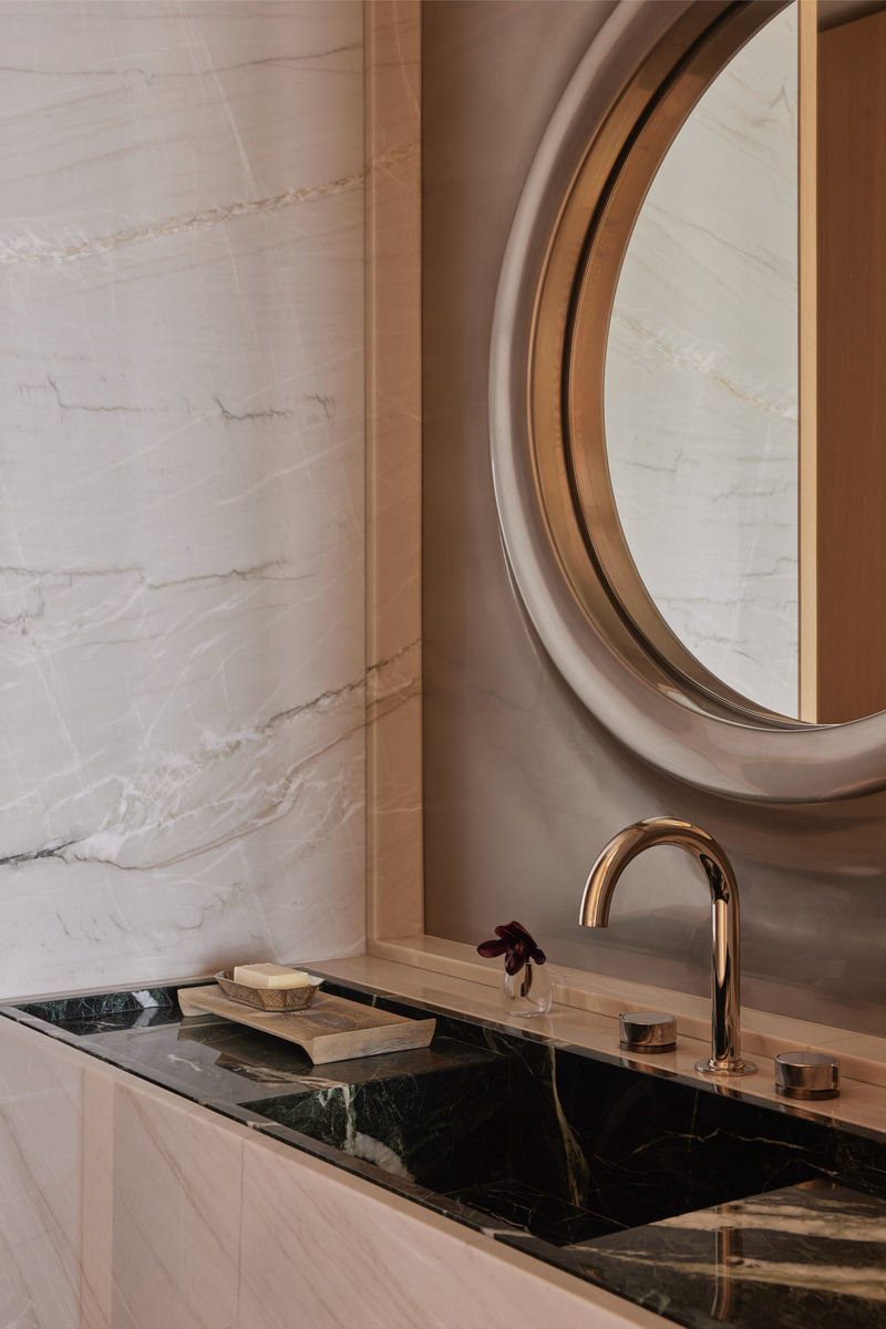 Press kit | 6087-07 - Press release | Study in Serenity: A Beachside Haven of Precision and Craftsmanship - Daniel Joseph Chenin - Residential Interior Design - A study in contrasts, the powder room vanity layers deep green marble against pale stone walls, its surface punctuated by a bronze faucet and circular mirror frame that echo the geometry of a ship’s porthole. Every detail, from the hand-carved soap tray to the single bloom, underscores a narrative of restraint, craft, and quiet luxury. - Photo credit: Tim Hirschmann