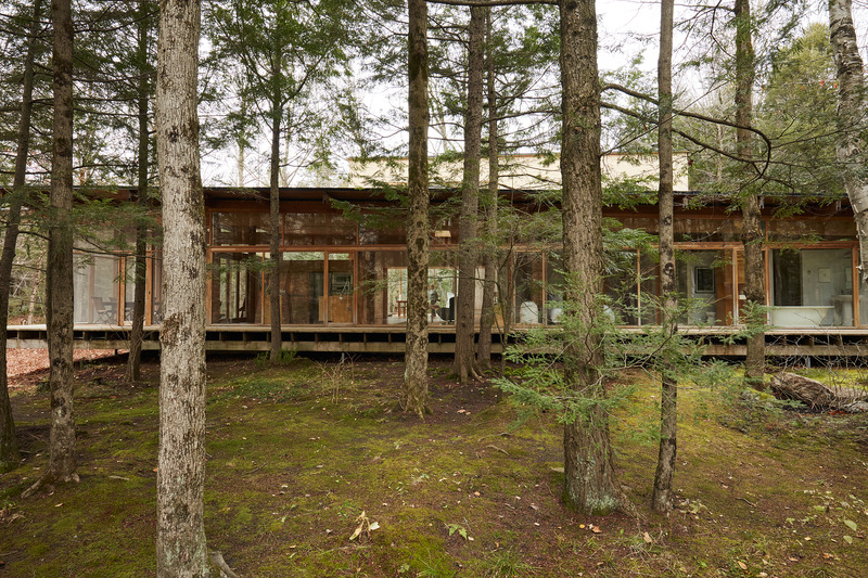 Press kit | 1640-01 - Press release | Territories and Landscapes Exhibition in Paris by Atelier Pierre Thibault - Atelier Pierre Thibault - Event + Exhibition - Résidence Les Abouts (2005), Saint-Edmond-de-Grantham, Centre du Québec<br>A woodland setting in the heart of the forest, this house is a place of communion where art and nature become one. Imagined for contemporary art collectors who dreamed of a spacious and warm home to display their works and accommodate their daily lives, the plan unfolds in two distinct parts separated by a central circulation. The first section, a single-storey building stretching over thirty meters, dialogues with the horizontal line of the river that circles the site. It seems to levitate above the ferns and mosses of the site, carefully maintained by the residents. In contrast, the second part rises on two floors, offering interior spaces that recall those of an art gallery. Everywhere, the openings frame views of the tree trunks, like landscape paintings that naturally dialog with the collected artworks. - Photo credit: Maxime Brouillet