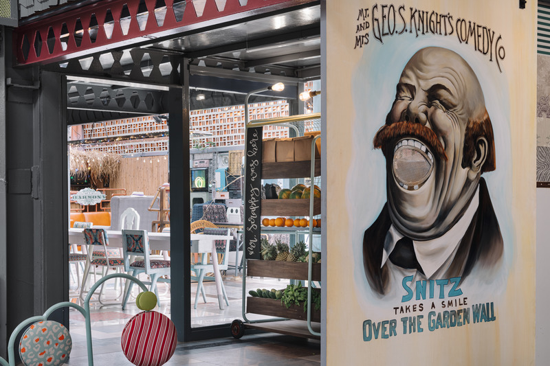 Press kit | 7002-01 - Press release | Big Top - Multitude Of Sins - Commercial Interior Design - <p>The entrance to the café is earmarked by a buffer zone that rests behind the veil of an artisanal barn door which seems to have been teleported through time from the confines of an underground speakeasy! Illustrated by Artist Shunnal Ligade, the Snitz’s merry mouth that splits wide open unable to contain his roaring laughter has been backed by a lining of perforated metal sheet. This ante space hosts a fleet of custom food carts that emulate the produce-to-plate concept in a new light with these ingenious contraptions. Finished in hues of pastel blue, yellow, and mint, these food carts are a humble homage to the ‘haathgaadis’ possessed by Indian vegetable and fruit hawkers. Curvilinear in their make, the carts exude an industrial nuance in the form of their filament bulbs fixtures, wooden crates, and tubular structure.</p> - Photo credit: Ishita Sitwala