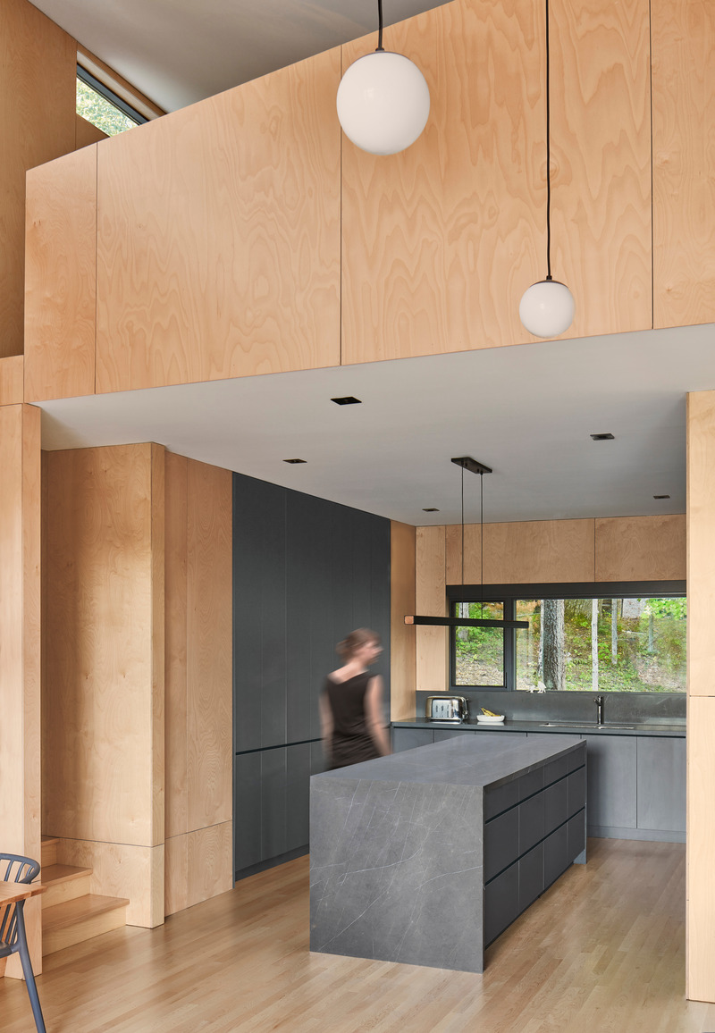 Dossier de presse | 1513-14 - Communiqué de presse | Catchacoma Cottage - Dubbeldam Architecture + Design - Architecture résidentielle - The kitchen, tucked beneath the loft, contrasts the warm hues of maple plywood with cool greys. It features Greystone counters, charcoal cabinetry, and a waterfall island. - Crédit photo : Riley Snelling